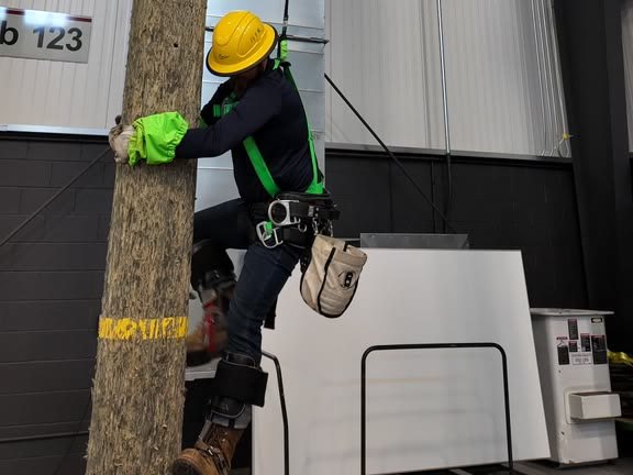 Trainee in a yellow hard hat and green safety harness climbing a pole inside the indoor training facility